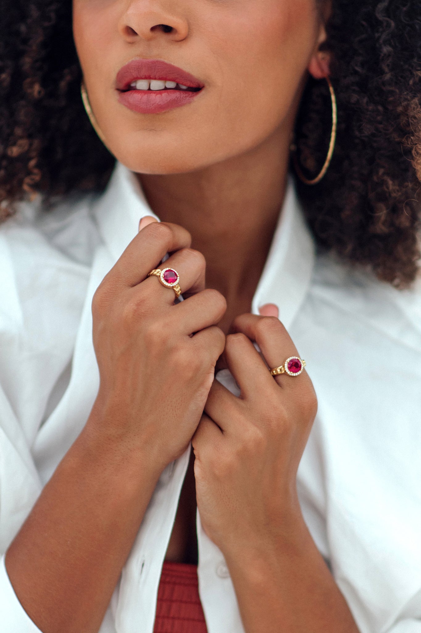 Woman wearing gold hoop earrings and rings with red gemstones, close-up of hands.